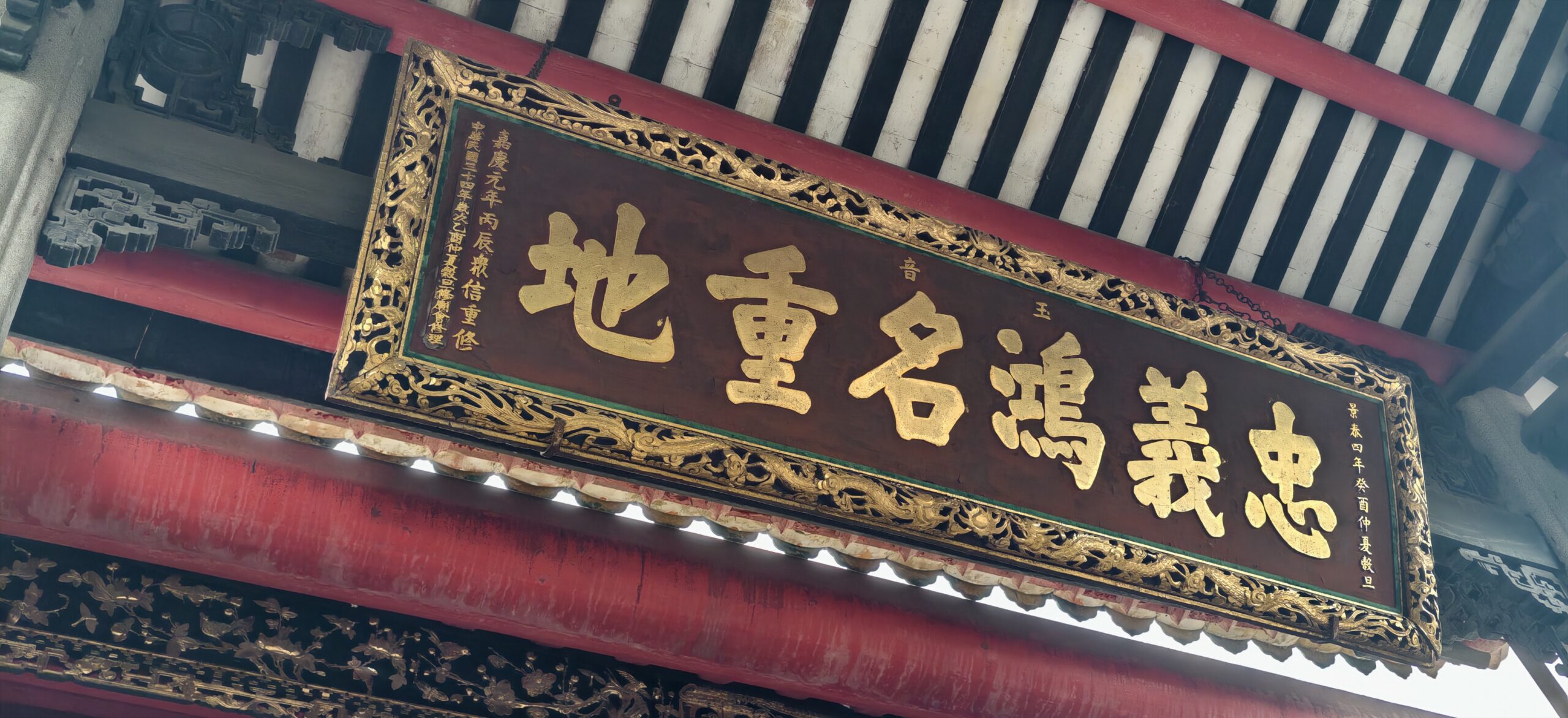 古建梁架间高悬忠义流芳名重地匾额，木雕细节密集。